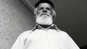 Un uomo anziano con barba bianca e cappello guarda verso il basso, in un'inquadratura dal basso in bianco e nero.