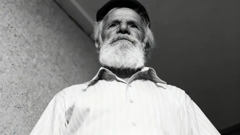Un uomo anziano con barba bianca e cappello guarda verso il basso, in un'inquadratura dal basso in bianco e nero.