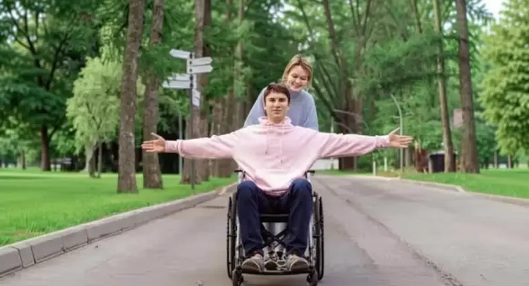 Una persona su sedia a rotelle con le braccia aperte e un'altra persona che lo spinge in un parco