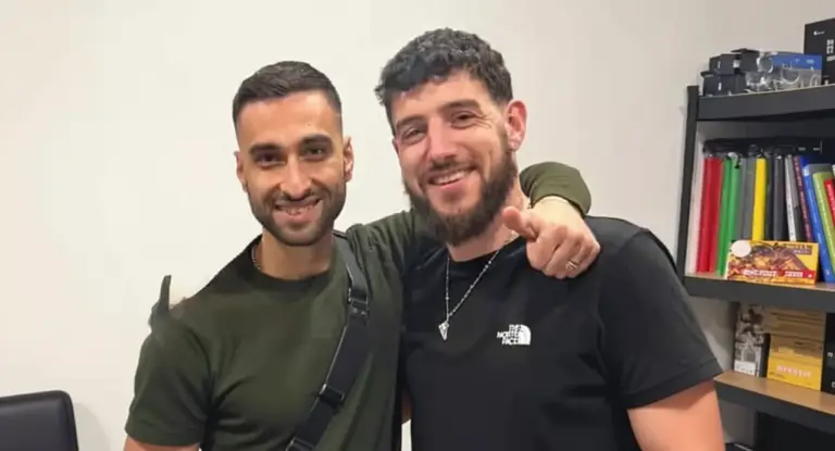 Due persone sorridenti posano insieme in un ufficio con scaffali pieni di libri e oggetti da collezione.