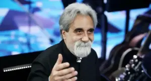 Un uomo con capelli e barba bianchi sorride in un ambiente televisivo con strumenti musicali sullo sfondo