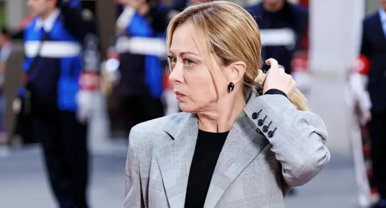 Una donna con espressione seria in un ambiente formale all'aperto, con persone in divisa sullo sfondo