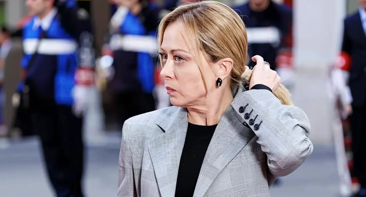 Una donna con espressione seria in un ambiente formale all'aperto, con persone in divisa sullo sfondo