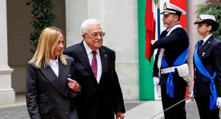 Due persone camminano affiancate in un cortile ufficiale con bandiere italiane sullo sfondo.