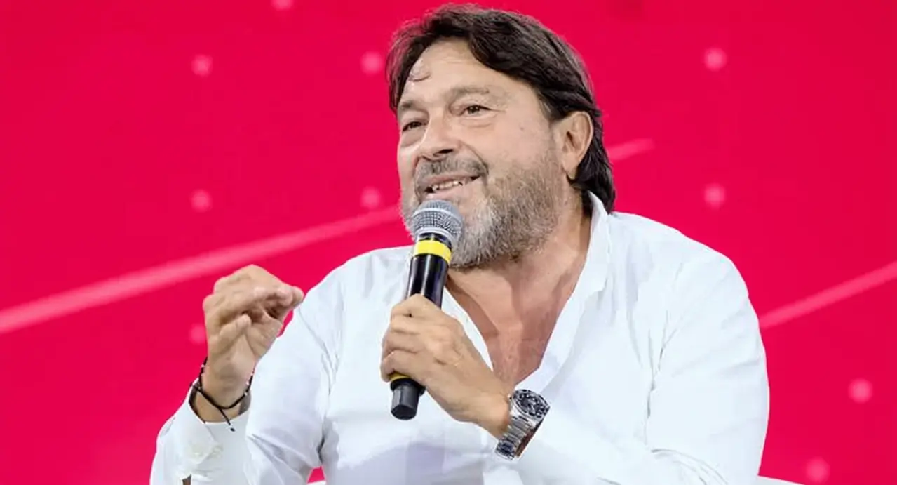 Un uomo con camicia bianca parla tenendo un microfono, su sfondo rosso