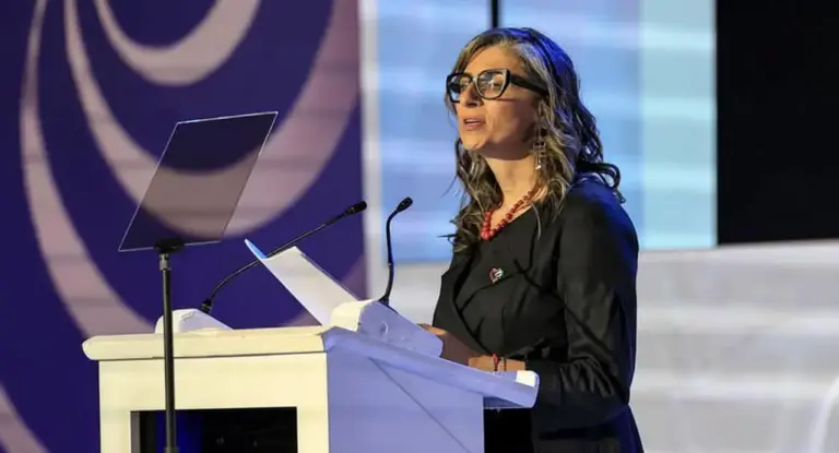 Una donna parla da un podio durante un evento pubblico, con microfoni e leggii davanti a sé.