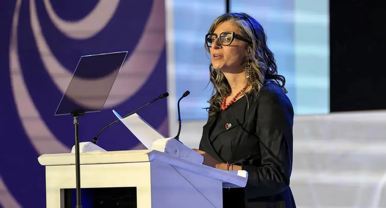 Una donna parla da un podio durante un evento pubblico, con microfoni e leggii davanti a sé.